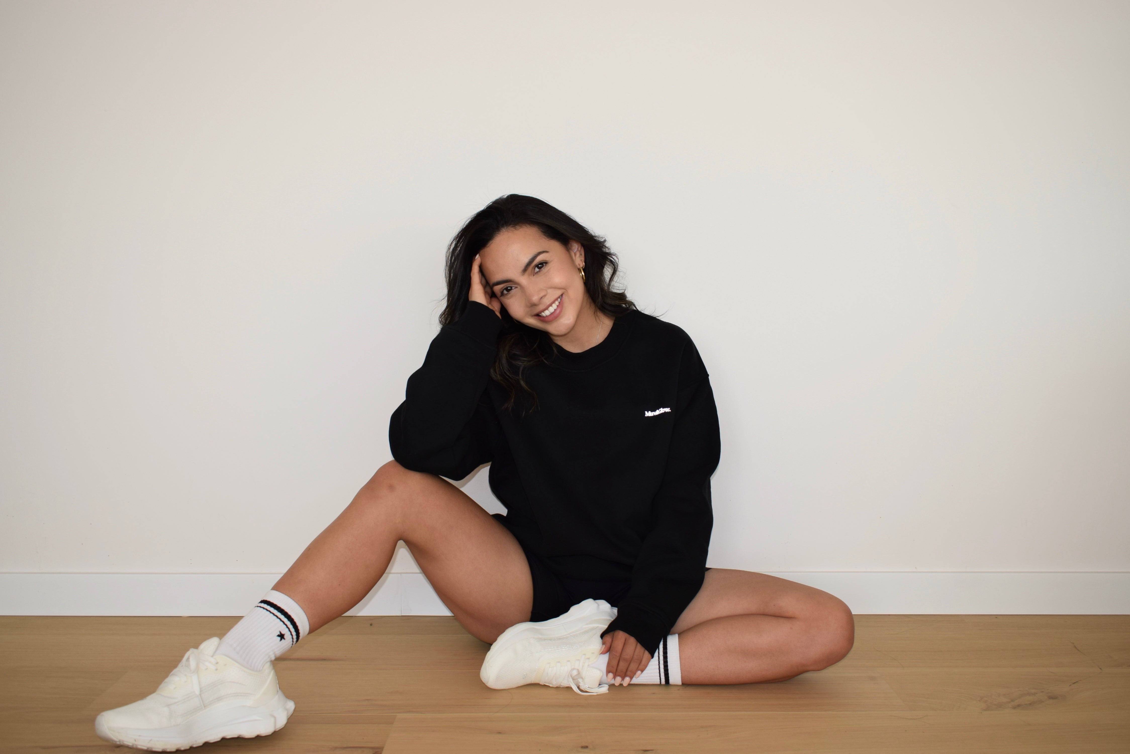 Modelo sentada en el piso usando el Essential Pullover negro de Muv&Glow, en ambiente neutro.