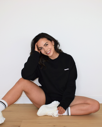 Vista relajada del Essential Pullover negro de Muv&Glow, modelo sentada con sudadera amplia y cómoda.
