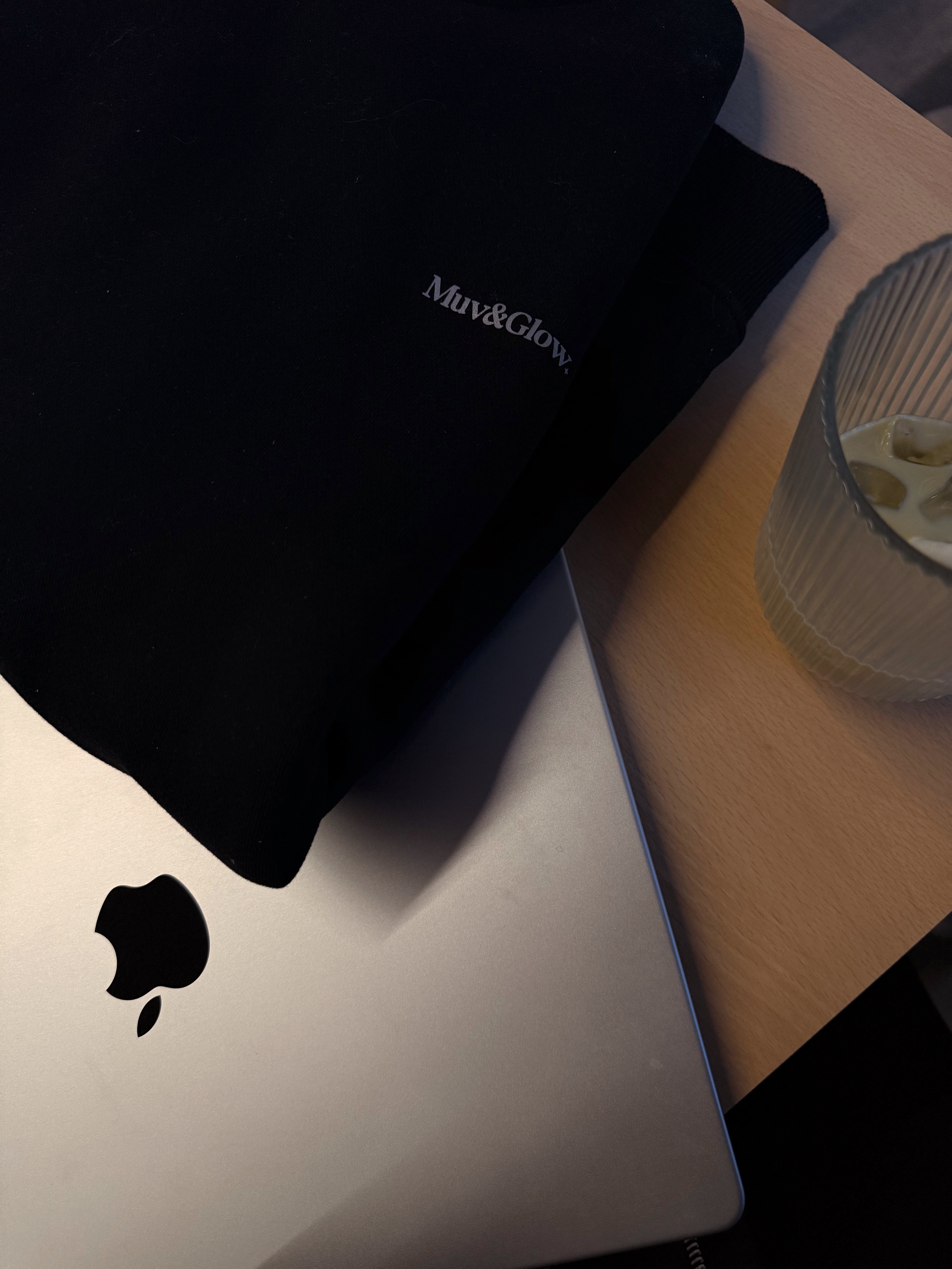 Detalle de Essential Pullover negro de Muv&Glow junto a un MacBook, ambiente de trabajo.
